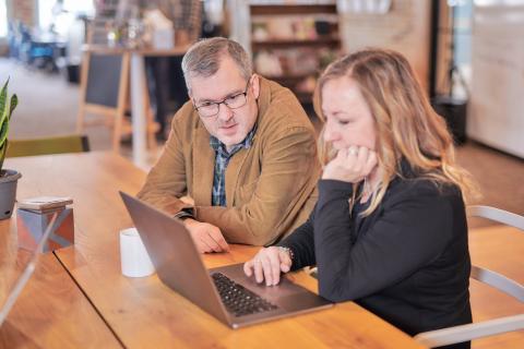 Jonathan sits next to coworker and helps solve a problem on laptop