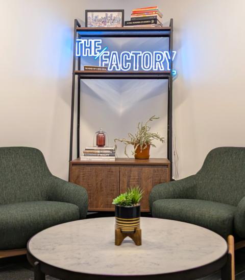 A glowing neon sign for The Factory on a shelf between two green chairs.