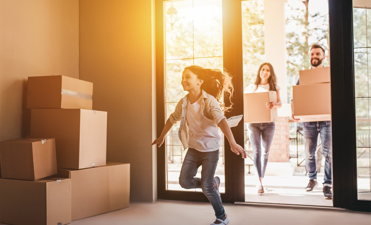 Girl runs happily into new home filled with boxes, followed by parents carrying boxes