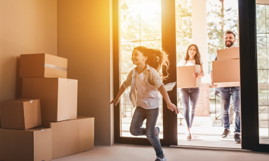 Girl runs happily into new home filled with boxes, followed by parents carrying boxes